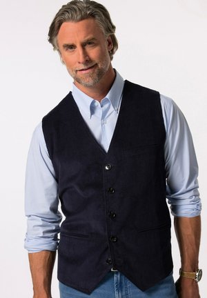 Gilet en velours côtelé bleu marine à cinq boutons, design en v, avec une poche sur le devant, porté au-dessus d'une chemise rayée bleu clair, associé avec un jean en denim.