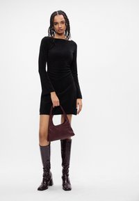 Abito nero a maniche lunghe con silhouette aderente, abbinato a stivali al ginocchio lucidi borgogna e una borsa coordinata. Design semplice e elegante.