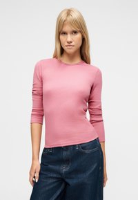 Γυναίκα που φοράει ροζ μακρυμάνικο ριμπ μπλουζάκι και σκούρα μπλε τζιν παντελόνια με φαρδύ πόδι, όρθια μπροστά σε σκέτο ανοιχτό γκρι φόντο.