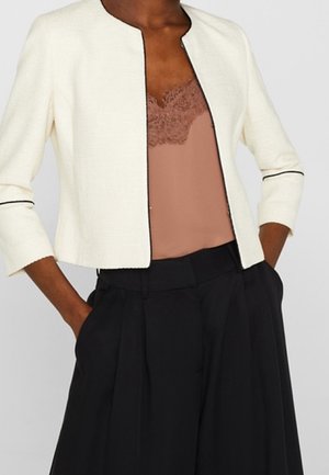 Femme portant une veste courte écrue ouverte sur le devant, un caraco mauve orné de dentelle, et un pantalon noir taille haute à jambes larges, les mains dans les poches.