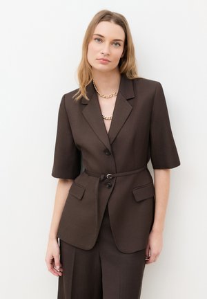 Kvinde iført en mørkebrun blazer med bælte og albuelange ærmer samt matchende bukser, accessoriseret med chunky guldkædehalskæder.