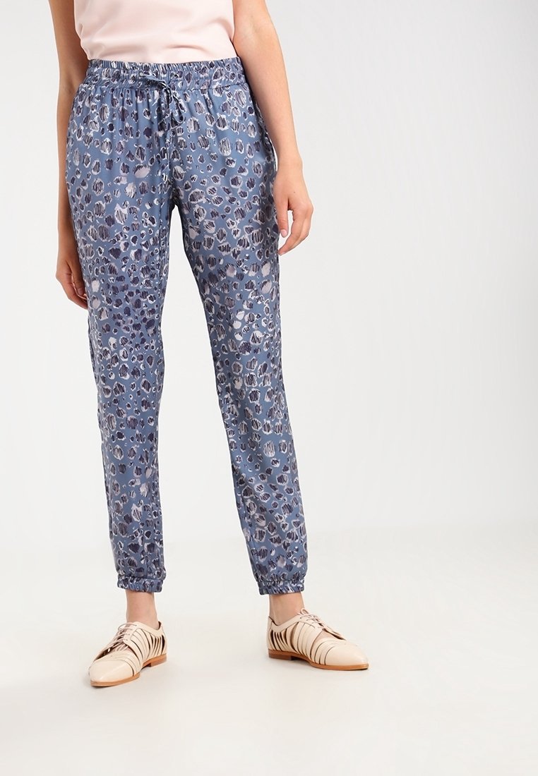 Personne portant un pantalon de jogging à taille élastique imprimé léopard bleu et des chaussures plates beiges à lacets, debout sur un fond uni.