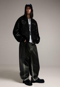 Fekete bombázó dzseki bőr ujjakkal, túlméretezett szürke farmernadrág és fekete fényes cipők. Egy fekete, logóval díszített sapka egészíti ki az összeállítást.