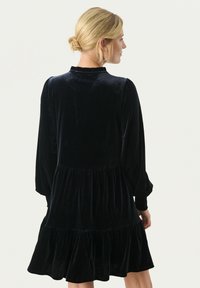 Rochie din catifea neagră cu mâneci lungi, tiv ondulat, corset ajustat și guler înalt. Textură moale și siluetă fluidă.