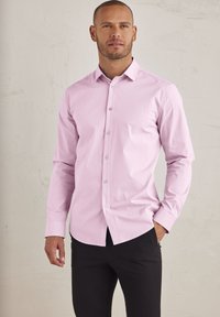 Helles rosa Hemd mit langen Ärmeln, klassischem Kragen und subtiler Textur. Kombiniert mit engen schwarzen Hosen, wodurch ein smart casual Look entsteht.