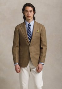 Man in een bruin blazer, lichtblauw overhemd, marineblauwe gestreepte das, witte broek en een gele pochet tegen een effen beige achtergrond.