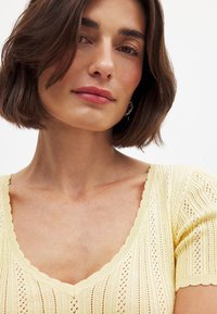 Geltona megztinė su V formos iškirpte, su tekstūruotu raštu ir banguotais krašteliais rankovėse ir apatiniame krašte. Minkšta, lengva medžiaga.