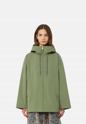 Donna che indossa una giacca verde oliva larga con cappuccio e tasche frontali, e una gonna a fantasia, in piedi contro uno sfondo semplice.