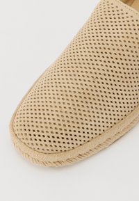 Scarpa beige slip-on con un design superiore perforato e dettagli in corda cuciti lungo la suola, che mostra una texture liscia.