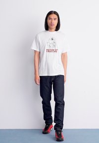 Witte katoenen t-shirt met rode "FAIRPLAY" graphic, gecombineerd met donkerblauwe jeans en zwart met rode accenten sneakers, staande op een lichtblauwe vloer.
