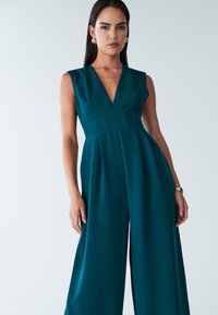 Jumpsuit sans manches teal avec un décolleté en V, des jambes larges et des coutures plissées. Texture lisse, design minimal, associé à des boucles d'oreilles en teinte dorée.