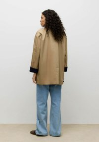 Chaqueta beige extragrande con puños azul oscuro, hecha de tela suave. Combinada con vaqueros de pierna ancha azul claro y zapatos marrones.