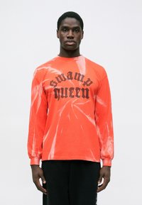 Muškarac u dugom, svijetlo narančastom majici s tie-dye uzorkom i crnim natpisom "swamp queen", nosi crne hlače i stoji protiv jednostavne pozadine.