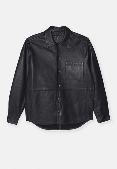 Svart läderskjorta med spetsig krage, framknappar, en bröstficka och en slät textur. Har en fåll som böjer sig något vid sidorna.