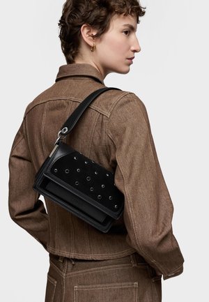 Mujer con chaqueta y pantalones de mezclilla marrones, llevando un bolso de hombro de cuero negro con ojetes metálicos sobre su espalda.