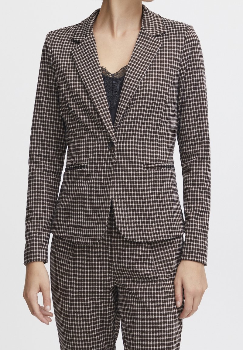 Femme portant un blazer marron et blanc à carreaux avec un pantalon assorti et un haut noir bordé de dentelle en dessous.