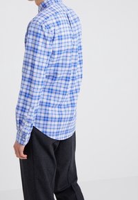 Man i blå och vit rutiga långärmad skjorta och mörkgrå byxor, vänd något för att visa skjortans baksida och sida.