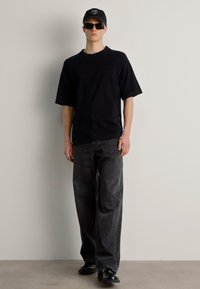 T-shirt nera oversize, jeans a gamba larga grigio scuro, occhiali da sole neri e cappello con testo bianco. Tessuto morbido, vestibilità casual.