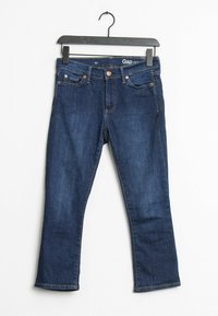 Jean en denim bleu foncé avec un design à cinq poches, coupe droite, rivets en cuivre et coutures beiges. Suspendu à un cintre en plastique noir.