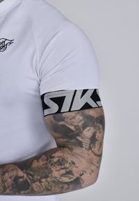Hvid atletisk skjorte med en tætsiddende design. Har et sort og gråt elastisk armbånd med teksten "STKS", samt detaljerede tatoveringer på armen.