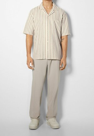 Hombre con camisa de botones de manga corta a rayas beige, pantalones beige claro y zapatillas blancas roto, de pie frente a un fondo blanco.