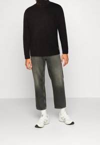 Schwarzer Langarm-Turtleneck, graue gerade geschnittene Jeans und weiße Sneaker mit schwarzen Akzenten, getragen von einer Person, die vor einem schlichten Hintergrund steht.