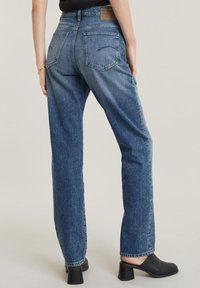 Plave traperice s visokim strukom, ravnim nogavicama i laganim izbljeđivanjem. Imaju dva stražnja džepa i našiveni natpis na pojasu.