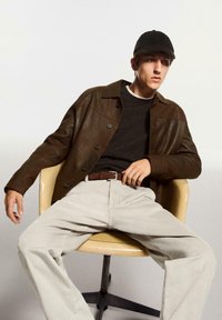 Veste en cuir marron, pull noir texturé, pantalon en velours côtelé beige, ceinture marron, casquette noire, assis sur une chaise couleur crème.