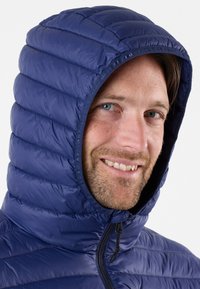 Marineblauwe puffy jas met een capuchon, voorzien van een geborduurde textuur en een ritssluiting aan de voorkant. Glad, glanzend materiaal en een aansluitende pasvorm rond het gezicht.
