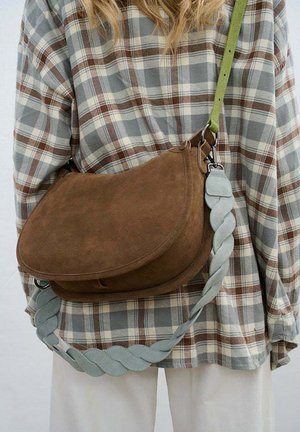 Hnědá semišová kabelka přes rameno s kulatým designem, vybavená propleteným světle modrým popruhem a kovovými doplňky. Nositelná přes károvanou košili.