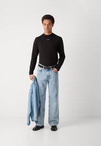 Musta pikad varrukad särk logoga, helesinised laiad teksad, hoides helesinist denim-jakki, mustad kingad, seistes halli tausta vastas.