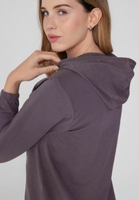 Sudadera con capucha en púrpura suave, que presenta una textura suave, mangas largas y un ajuste casual. Incluye un bolsillo frontal y cordones ajustables.