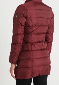 Abrigo acolchado burdeos con diseño acolchado, cuello alto, mangas largas y bolsillo lateral con cremallera. Textura suave y lisa, largo hasta la rodilla.