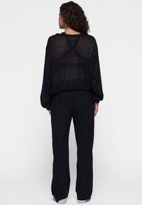 Schwarzer transparenter Pullover mit langen Ärmeln und lockerer Passform. Kombiniert mit schwarzen Hosen. Auffällige Kreuz-Detailverarbeitung im Rücken.