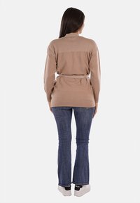 Cardigan belted color marrone chiaro con texture a costine sulle maniche, abbinato a jeans svasati blu scuro e sneakers bianche.