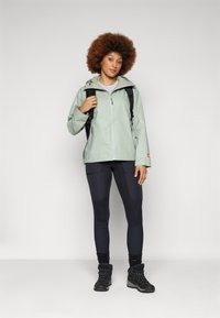 Veste imperméable vert clair avec capuche, associée à des leggings noirs avec des patchs texturés et des bottes de randonnée noires.