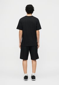 Άνδρας με σγουρά μαλλιά στέκεται με την πλάτη στραμμένη φοράει απλό μαύρο μπλουζάκι, μαύρα σορτς μέχρι το γόνατο, λευκές κάλτσες και μαύρα αθλητικά παπούτσια.