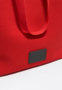 Rode textuur tote bag met stevig materiaal, stoffen handvatten en een zwart logo label met verhoogde tekst.