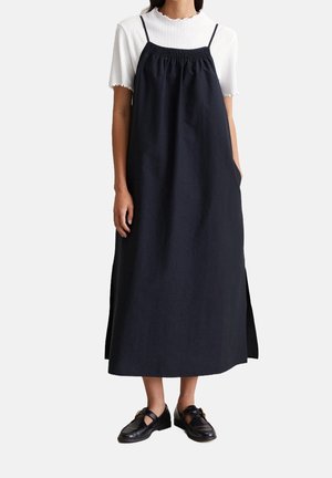 Nainen, jolla on päällään musta hihaton midimekko valkoisen ribbineuloksisen lyhythihaisen paidan päällä ja mustat loafersit, seisoo valkoista taustaa vasten.