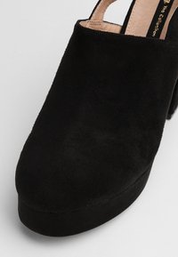 Chaussure à plateforme en suède noir avec un bout arrondi et un dos ouvert, présentant une texture lisse et un talon épais pour plus de hauteur.