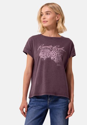 Femme blonde portant un t-shirt violet ample avec un graphisme de tête de léopard et le texte "Rewrite Rules", associé à un jean bleu.