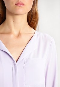 Blusa de color púrpura claro con una textura suave, que presenta un escote en forma de V y un bolsillo frontal. La tela cae suavemente sobre los hombros.