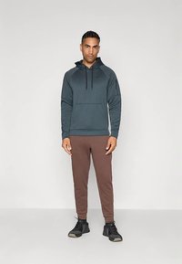 Sötét türkiz kapucnis pulóver elülső zsebbel, barna slim-fit melegítőnadrággal és fekete sportcipővel párosítva.