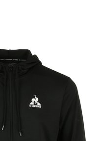 Zwarte trainingjack met ritssluiting en capuchon, met een wit logo op de borst en een gladde, elegante stoftextuur.