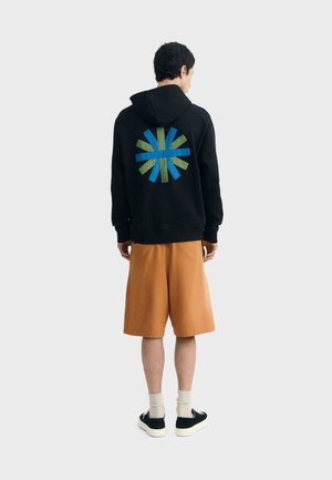 Person wearing black hoodie with blue and green abstract design on back, tan knee-length shorts, white socks, and black sneakers, standing facing away.