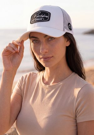 Frau mit langen dunklen Haaren, die ein beiges Hemd und eine weiße Blackskies Baseballkappe trägt, richtet die Kappe an einem Strand bei Sonnenuntergang.