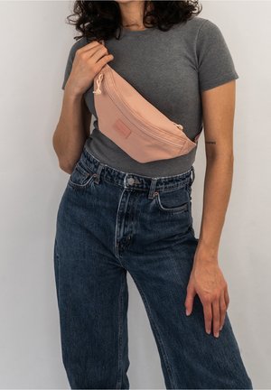 Persona que lleva una camiseta gris y vaqueros azules, sosteniendo un bolso bandolera rosa claro cruzado sobre el pecho contra un fondo liso.