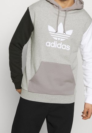 Mężczyzna noszący kolorowy kaptur Adidas z czarnym lewym rękawem, białym prawym rękawem, szarą częścią główną i przednią kieszenią, stojący na jednolitym tle.