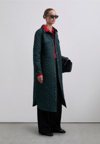 Abrigo exterior acolchado verde con botones a presión, grandes bolsillos, cuello rojo, combinado con pantalones negros y un bolso estructurado negro.