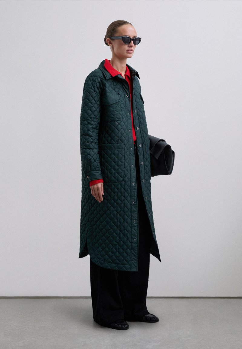 Abrigo exterior acolchado verde con botones a presión, grandes bolsillos, cuello rojo, combinado con pantalones negros y un bolso estructurado negro.